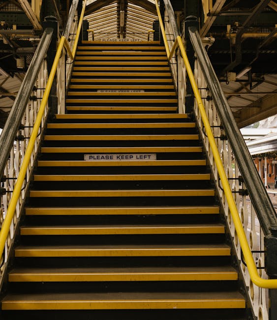 A set of industrial metal stairs with yellow safety rails on each side, leading up to an elevated platform within a warehouse or loading dock area. The stairs are covered with non-slip yellow and black striped tread covers and have a “PLEASE KEEP LEFT” sign attached to the middle step, indicating a safety instruction for movement and traffic flow. The surrounding environment includes structural metal beams, pipes, and underfloor lighting, suggesting a utilitarian setting for home relocation or furniture transport. The open space and visible lighting imply good visibility for loading and unloading operations, supporting efficient moving processes managed by companies like removal companies West Kensington. The image captures the staircase in a professional, industrial context, highlighting typical access difficulties during house removals and the importance of careful planning in narrow or complex staircases for safe and effective furniture transport.