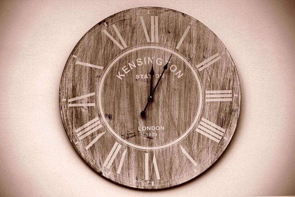 A round wall clock with a rustic, distressed wood finish face, displaying Roman numerals in white indicating the time as 12:02. The clock features the words 'KENSINGTON STATION', 'LONDON', and the year '1809' printed in the centre. It is mounted on a plain, light-colored wall, with no other objects visible nearby. This clock may be part of an interior setting used for timekeeping during home relocation or furniture transport planning, which relates to removals services offered by removalcompanieswestkensington.co.uk.