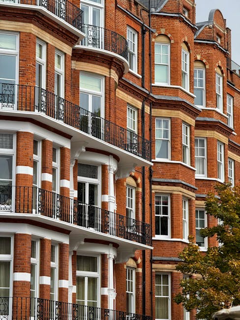 A multi-storey red brick residential building with white decorative bands and black metal balconies overlooking large windows. The structure features curved bay windows and traditional architectural details typical of London apartment buildings. There are trees with green foliage partly visible in the lower right corner, and the clear sky is reflected in some of the windows. This building is situated in an urban area suitable for house removals and home relocation services, as provided by removal companies in West Kensington. The image captures the exterior façade, illustrating the type of property involved in furniture transport and moving logistics, which often requires careful planning for narrow access points, as highlighted in the Earl Court flat removals checklist for narrow access.