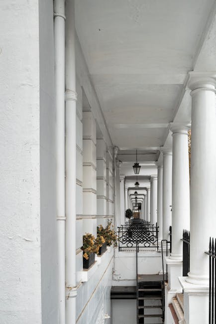 A view of a narrow white-painted residential balcony corridor in Earl Court, with evenly spaced white columns supporting the ceiling. Several small potted plants with dried or yellowing foliage are arranged along the edge of the corridor, placed on a ledge. A black wrought iron fence with decorative patterns runs along the edge of the balcony, separating it from the stairwell below. In the distance, there are two black outdoor lanterns mounted to the ceiling, hanging along the corridor. The environment is well-lit by natural lighting, with sunlight creating soft shadows on the walls. This scene depicts a typical residential exterior space suitable for a home relocation project, with an emphasis on narrow access characteristics relevant to the removals and furniture transport process, as showcased by the tight, step-limited pathway.