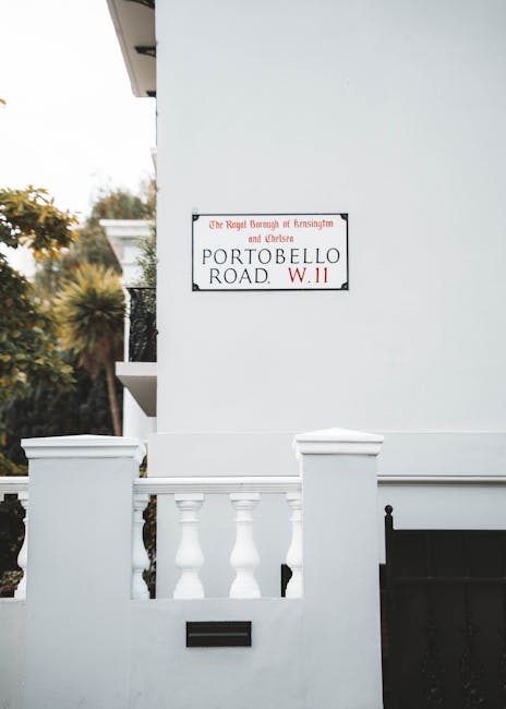 A white residential building's exterior wall features a street sign for Portobello Road, positioned on the upper section of the wall beside a small balcony with decorative white balustrades. The building has a clean, smooth finish, and the sign indicates the jurisdiction of The Royal Borough of Kensington and Chelsea, with a red W.11 postal code. The lower part of the wall includes a black mailbox and a black metal gate, possibly leading to a driveway or entrance area. The scene is captured during daylight with soft natural lighting, and in the background, lush green trees are visible, suggesting a peaceful neighbourhood setting. This visual is relevant to house removals or home relocation services, illustrating the typical exterior environment encountered during property moving logistics or packing and moving procedures undertaken by companies like [COMPANY_NAME] for clients in the Kensington area.