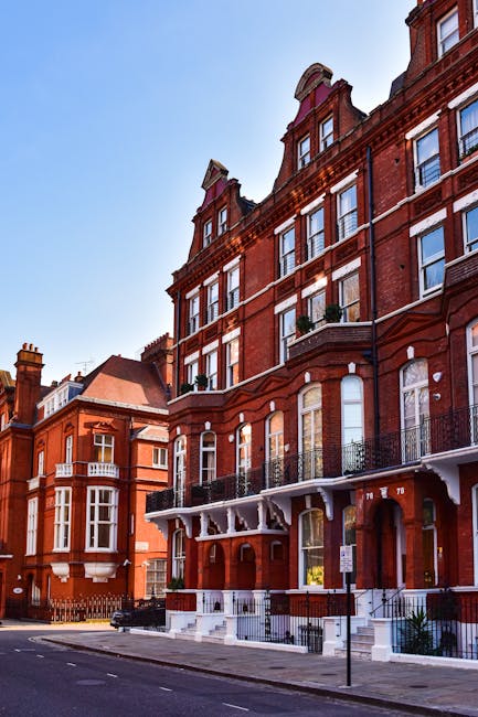 Image of a red brick residential building with multiple floors, large windows, and decorative iron balconies on the upper levels. The building features a prominent entrance with white steps leading to a doorway, adjacent to a pavement where a moving trolley loaded with cardboard boxes and wrapped furniture is being used for home relocation. The scene shows a clear blue sky, and other similar buildings line the street. Nearby, a van is parked, and the loading process appears to be underway, supported by moving blankets, straps, and a trolley for furniture transport, reflecting typical packing and moving procedures undertaken by professional removals services such as those offered by [COMPANY_NAME]. The image captures the exterior environment during daylight, illustrating a typical urban setting for house removals in Kensington, with attention to detail in packing materials and the logistics of loading items for a home relocation.