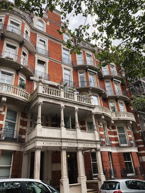 A white residential building's exterior wall features a street sign for Portobello Road, positioned on the upper section of the wall beside a small balcony with decorative white balustrades. The building has a clean, smooth finish, and the sign indicates the jurisdiction of The Royal Borough of Kensington and Chelsea, with a red W.11 postal code. The lower part of the wall includes a black mailbox and a black metal gate, possibly leading to a driveway or entrance area. The scene is captured during daylight with soft natural lighting, and in the background, lush green trees are visible, suggesting a peaceful neighbourhood setting. This visual is relevant to house removals or home relocation services, illustrating the typical exterior environment encountered during property moving logistics or packing and moving procedures undertaken by companies like [COMPANY_NAME] for clients in the Kensington area.