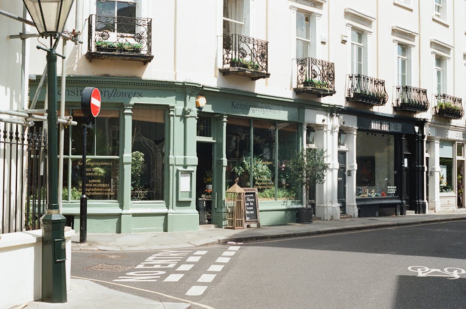 A street view showing a row of traditional white and pastel green buildings with large front windows, some featuring decorative wrought iron balconies adorned with potted plants. The ground floor of the building on the left is a flower shop, with signage reading 'Kensington Flowers' and displaying floral arrangements inside through the glass window. Outside the shop, a small chalkboard sign and potted plants are positioned near the sidewalk. In front of the buildings, there is a paved street with a bicycle lane marked on the road surface and a 'no entry' road sign mounted on a pole near the corner. Visible are a green bollard and a metal fence on the left side. The scene is outdoors during daylight with natural light, capturing the ambience of a typical West Kensington residential and retail area, suitable for highlighting home relocation or furniture transport aspects involving street access to the properties.
