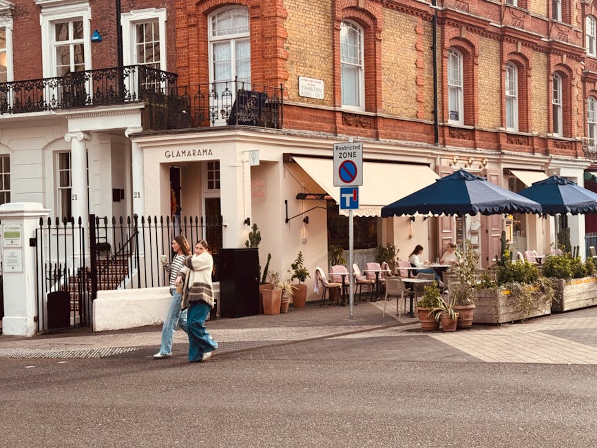 A street-facing view of a property at High Street Kensington, showing a brick building with large multi-pane windows and ornate iron balcony railings. In front of the building, there is a cafe or restaurant with an outdoor seating area that features pink chairs, several tables, and large navy blue umbrellas providing shade. Potted plants and greenery decorate the sidewalk terrace. Two women, one carrying a bag, are walking on the pavement near a black gate that leads into the property, which has a white facade with the sign 'Glamarama' above the entrance. A street sign indicating a restricted zone and a no-parking zone is visible near a lamppost. The scene is taken during daylight hours with clear lighting showing the organized outdoor dining area and urban residential environment, reflecting a typical London neighbourhood linked to local moving or relocation activities in west Kensington.
