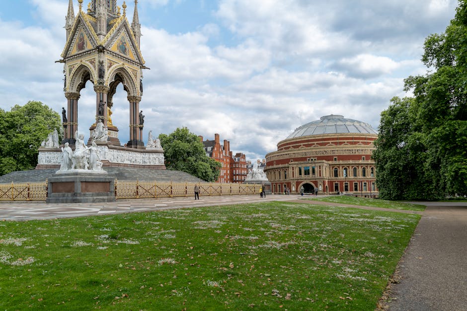 A wide view of a public park area in West Kensington, showing a large, ornate, multi-tiered monument with intricate sculptures and decorative details, located on a paved circular area surrounded by low fencing. To the right, there is a large, historic, circular building with a domed roof and arched windows, constructed with brick and stone materials. Lush green trees frame the scene on both sides, and a paved walkway extends from the foreground towards the monument and building. Some pedestrians are visible walking in the distance, and the overcast sky above has light clouds. The scene highlights an outdoor urban space that could be part of a home relocation or moving process involving transportation of items or equipment near notable city landmarks, as associated with professional removals services in West Kensington.