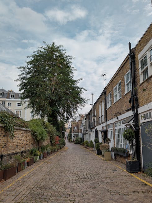 An empty residential cobblestone alleyway in West Kensington, with brick terraced houses on the right side, featuring large sash windows and white window frames. Potted plants and small shrubs line the base of the buildings and along the pavement on the left, which is bordered by a low brick wall with greenery. A tall tree with leafy branches extends above the alley, partially obscuring the sky, which is filled with scattered clouds. The setting appears calm and quiet, suitable for home relocation or furniture transport purposes. This image is relevant for content about Kensington parking permits for removals and council rules, illustrating the typical environment where removals services like those from [COMPANY_NAME] operate during house moves in the area.