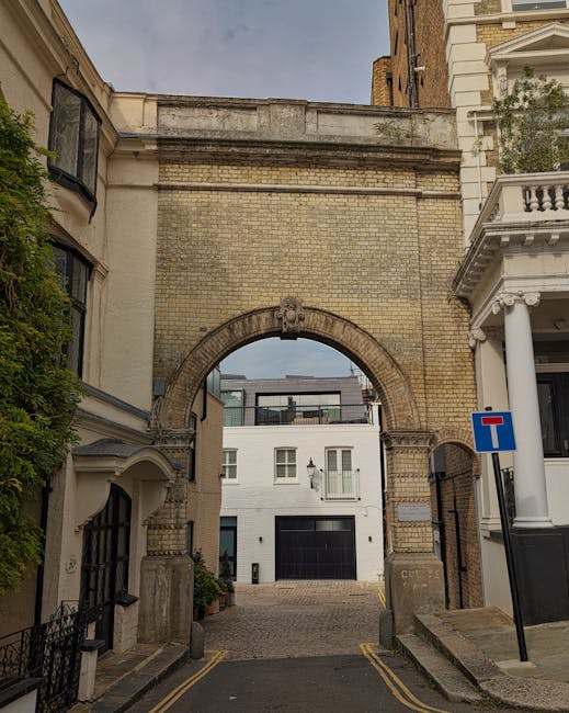 A narrow street leading to a gated residential property in West Kensington, with a large brick archway passing over the road, forming an entrance to the courtyard or driveway area. On the right side, a white building with classical columns and decorative trim is visible, along with a blue 'no entry' traffic sign. The pavement is paved with cobblestones, and the street is flanked by traditional townhouses with varying architectural styles. The scene is photographed in natural daylight, capturing the urban environment typical of house removals and furniture transport logistics, with an emphasis on the historic brickwork and building facades related to house moving services on the PAGE_TITLE page.