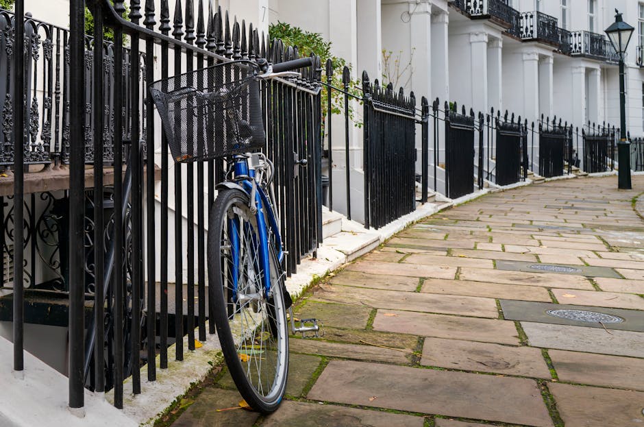 A blue bicycle with a black front basket is locked to a black wrought iron fence adjacent to a white building with classical architectural features, including white columns and decorative railings, on a paved sidewalk in West Kensington. The pavement consists of rectangular stone slabs with visible joints and some patches of moss or dirt along the edges. Several manhole covers are embedded in the sidewalk. The scene is well-lit, suggesting daytime, with the bicycle positioned close to a flight of steps leading up to the building's entrance. In the background, more black fences and additional white building facades with similar architectural details are visible, indicating a residential area. This setting illustrates typical urban residential surroundings where house removals or transport of furniture might be coordinated, with [COMPANY_NAME] often involved in such home relocation or moving logistics processes.