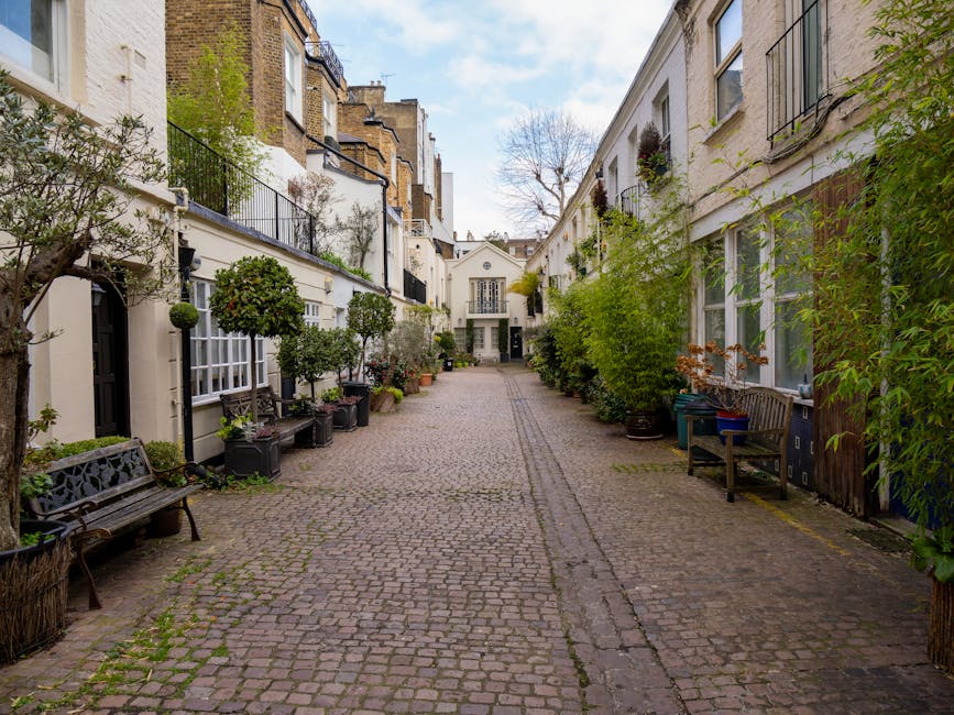 A cobbled courtyard in West Kensington with residential buildings on both sides, featuring white facades and large windows adorned with flower boxes. Along the pavement, there are wooden benches, potted plants, and small trees in containers, creating a tidy outdoor space. In the background, a white building with a central doorway and symmetrical windows can be seen, indicating a typical residential home. The scene appears to be during daytime with natural light illuminating the area. The image supports content related to house removals and moving services by illustrating an exterior location where furniture and boxes may be loaded or unloaded during a home relocation. The setting aligns with the city environment and moving logistics, emphasizing outdoor preparation and access points relevant to planning for pavement and skip licences, as advised for West Kensington moves by removal companies such as [COMPANY_NAME].