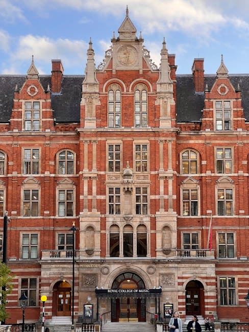 The image shows the exterior facade of the Royal College of Music in West Kensington, a historic red brick and stone building with ornate architectural details, including pointed gables, arched windows, and decorative stone carvings. In front of the building, there are several cardboard boxes and wrapped furniture pieces positioned on the pavement, indicating a home relocation or moving activity. A person is seen carrying a box towards a van parked nearby, which is partially visible in the lower right corner, with the back doors open, suggesting loading or unloading for a house move. The scene is set during daylight hours, with a clear sky and a few pedestrians walking along the street. This setting illustrates typical stages of furniture transport and packing during a professional removals process, supported by [COMPANY_NAME], natural to moving logistics within the context of local house removals service near Kensington.