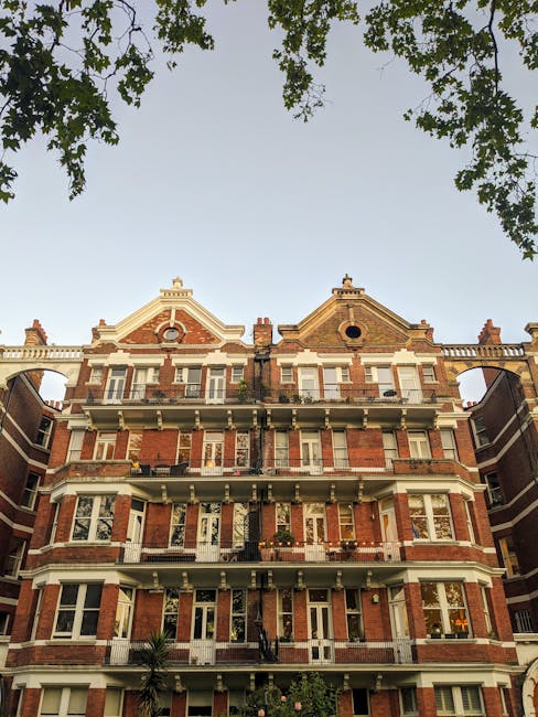 The image displays a multi-storey, red-brick residential building with classic architectural features, including decorative white stone window frames, balconies with black metal railings, and dormer windows on the top floor. Several cables are visible hanging from the second-floor balcony, and the building is framed by green tree branches at the top of the image, suggesting the photograph was taken from street level on a clear day with ample natural light. This setting aligns with house removals and relocation services in the West Kensington area, as shown on the domain removalcompanieswestkensington.co.uk, and could relate to logistical planning for furniture transport or packing for home relocation.