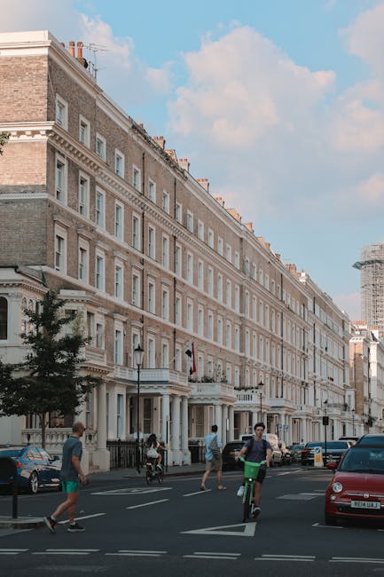 Image showing a row of multi-storey Victorian terraced townhouses with cream-colored facades, decorative cornices, and large sash windows, located on a busy street in West Kensington. The building features a small front garden area with black iron railings and a few outdoor lamps. In the foreground, pedestrians are walking along the pavement, and a cyclist is riding on the road, which is lined with parked cars, including a red vehicle. The scene is set during daytime with clear blue skies and some scattered clouds, illustrating an urban environment suitable for house removals and home relocation services, such as those provided by [COMPANY_NAME]. The street's atmosphere suggests an active residential area within West Kensington, where moving logistics and furniture transport may take place, supported by professional removals specialists.