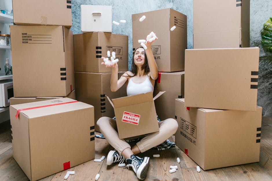 A woman sitting on the floor inside a room surrounded by numerous cardboard moving boxes of varying sizes, some sealed with red packing tape and others open with flaps folded back. She is looking up and smiling while tossing packing foam peanuts into the air, which are scattered around her on the wooden floor. Behind her are more boxes stacked against a textured light blue wall, and a doorway is partially visible with additional boxes and packing materials inside. The scene suggests she is engaged in home relocation, packing and moving process, with the boxes prepared for transport. The environment is well-lit, likely by natural or interior lighting, and the image reflects logistics associated with house removals, possibly coordinated by a professional moving service such as [COMPANY_NAME]. The overall setting emphasizes the packing phase of a furniture transport or home relocation, with packing materials and the woman’s cheerful demeanor indicating an efficient moving operation.