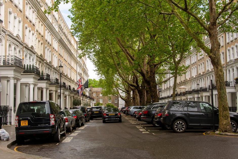A street scene in West Kensington with rows of elegant white terraced houses featuring black wrought iron balconies, large sash windows, and decorative molding. The street is lined with mature trees with lush green foliage, creating a canopy above the parked vehicles. Several cars, including black, silver, and grey models, are parked on both sides of the narrow road, some extending into designated parking bays marked by white lines. The pavement is wet, suggesting recent rain, and the environment appears calm and residential. Occasionally visible are black bollards and street lamps, with a Union Jack flag hanging from one of the houses. This setting provides a typical backdrop for home relocation or furniture transport activities, where moving companies like [COMPANY_NAME] might operate to assist with packing, loading, and transportation of household items amidst a peaceful, characterful neighbourhood.
