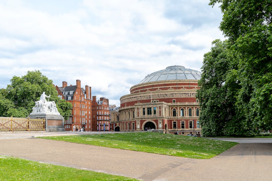 A wide view of a historic urban park area showing a large, rounded classical concert hall with a domed roof and detailed brickwork in the background, adjacent to several red-brick residential buildings with chimneys. In the foreground, a paved pathway runs through a grassy lawn, with the edge lined by tall, leafy green trees on the right and a statue on a pedestal on the left side of the image. The statue depicts multiple figures and is positioned on a decorative base with surrounding fencing. The scene is captured during daytime with partly cloudy skies, providing natural light that highlights the architecture and greenery, emphasizing the outdoor setting typical for house removals and moving logistics near iconic landmarks.